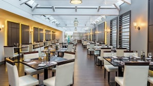 A spacious restaurant with high ceilings, skylights, and modern lighting fixtures. Tables are neatly arranged with chairs, and the interior features a clean, contemporary design.