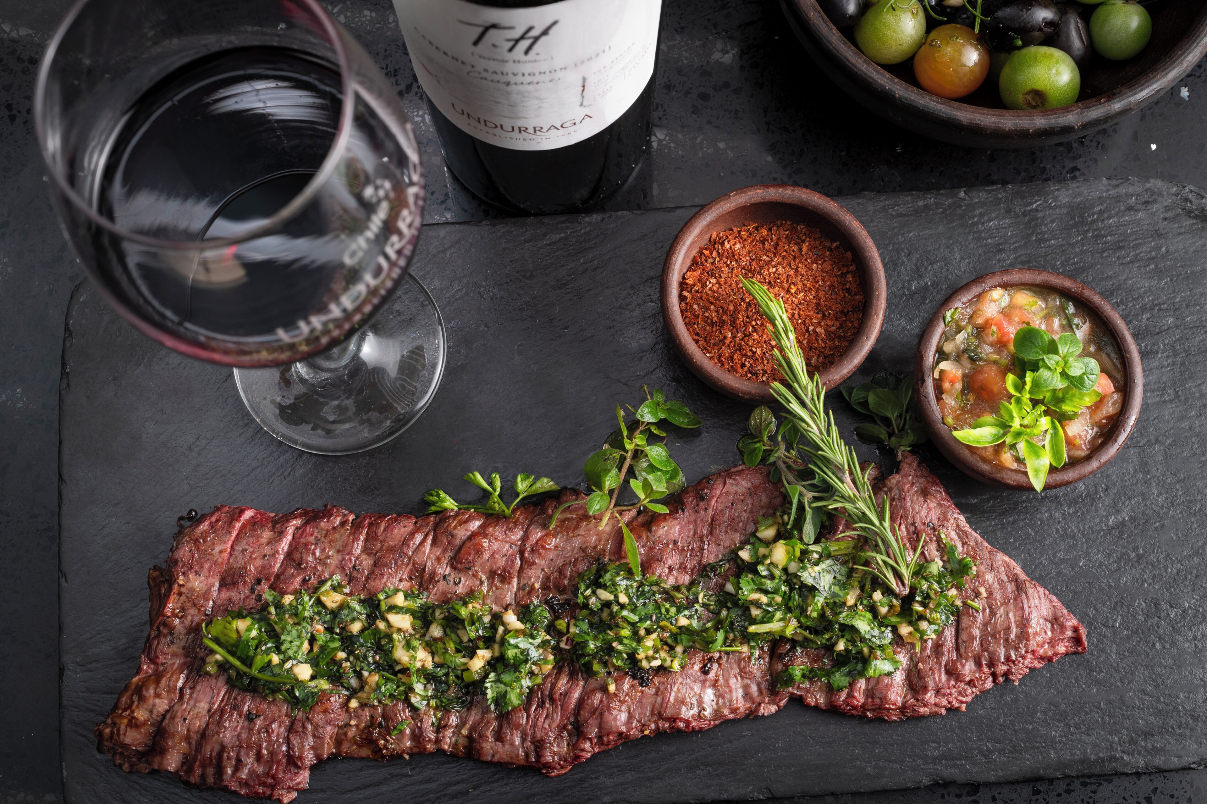Un bife con hierbas, una copa de vino tinto y dos recipientes pequeños con especias y salsa sobre una superficie de pizarra.