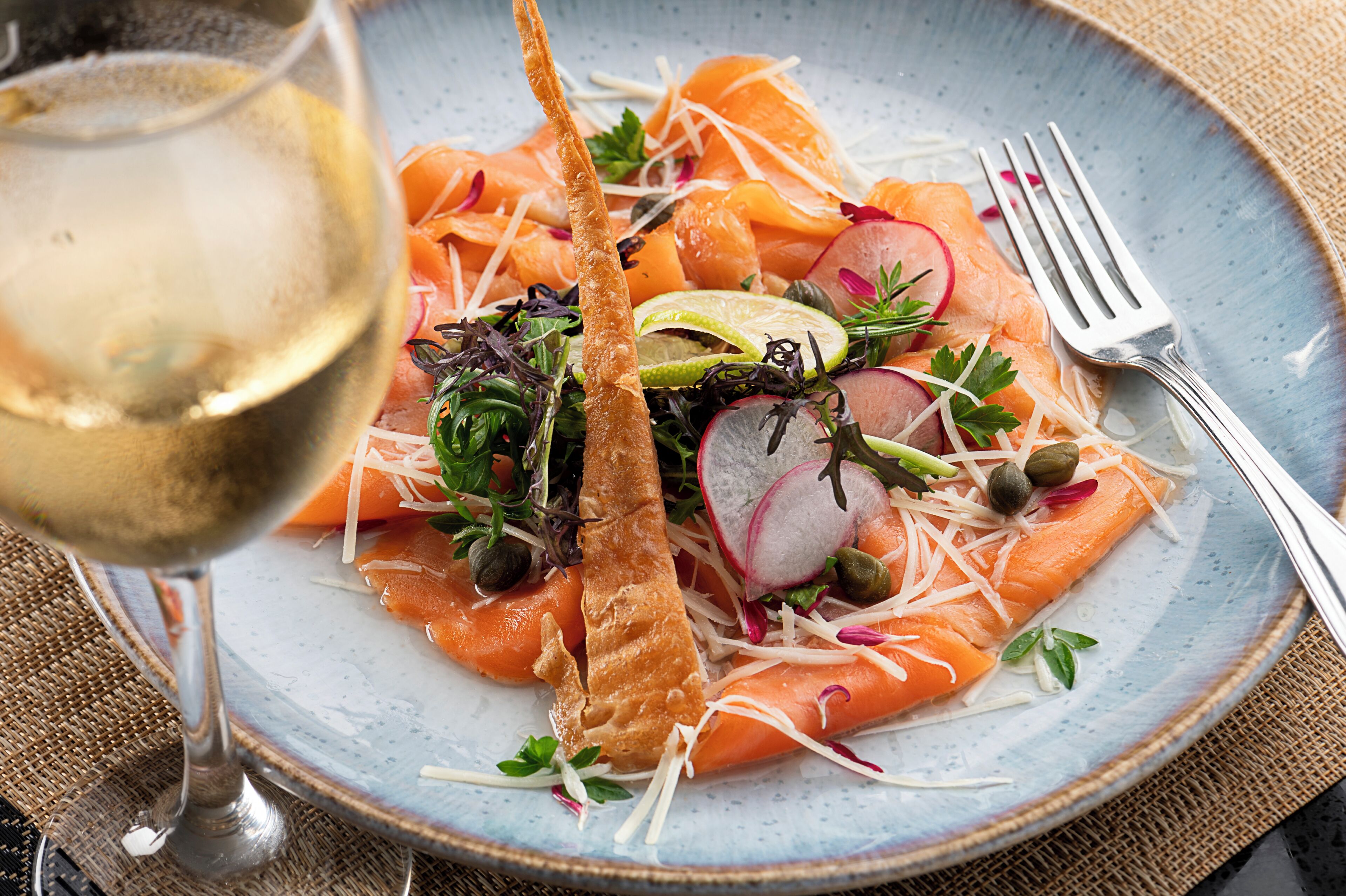 A plate of salmon salad with radishes, capers, and a slice of bread, accompanied by a glass of white wine.