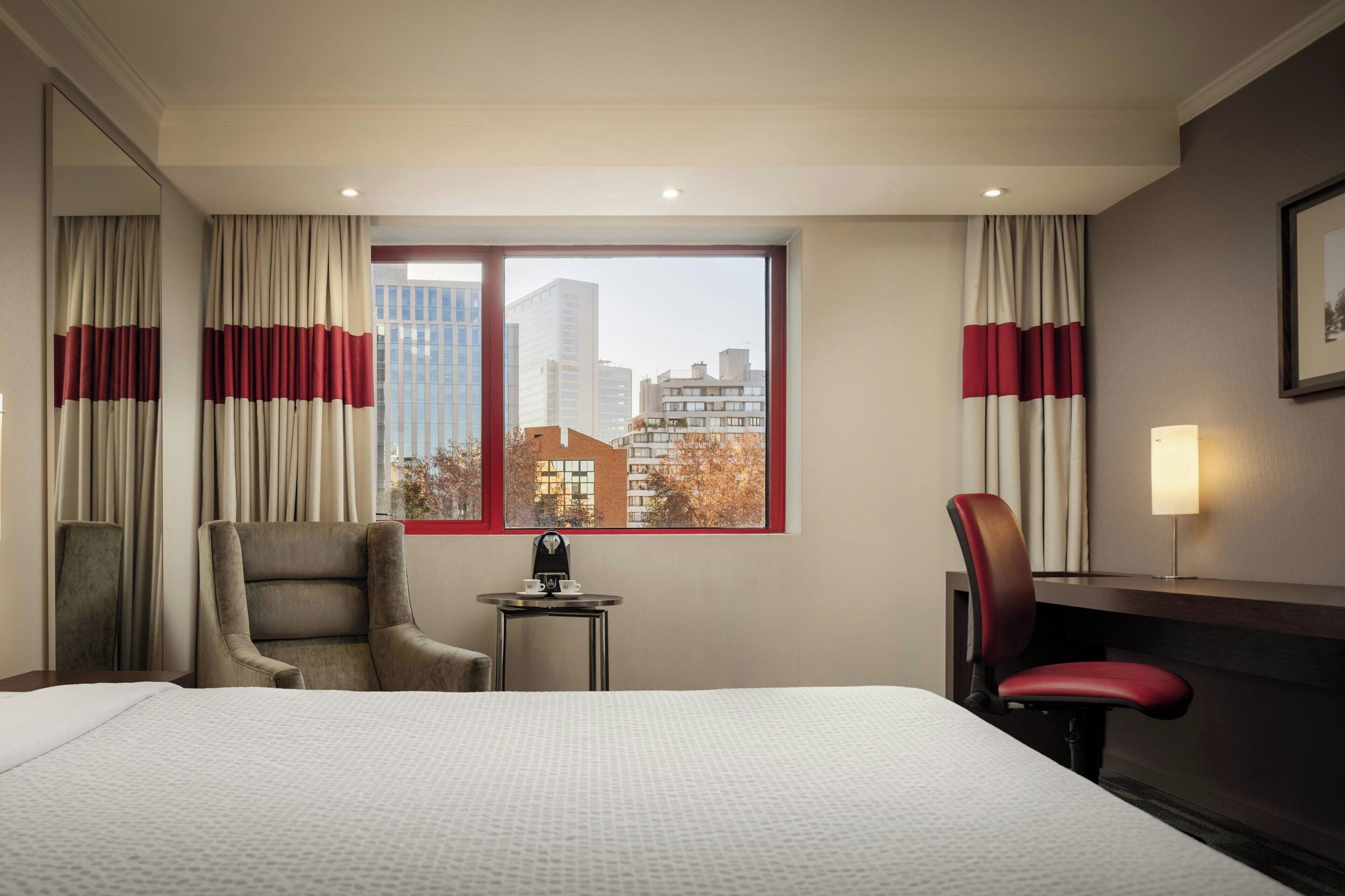A hotel room with a large bed, two chairs, a small table with a tea set, and a view of city buildings through a window with red curtains.