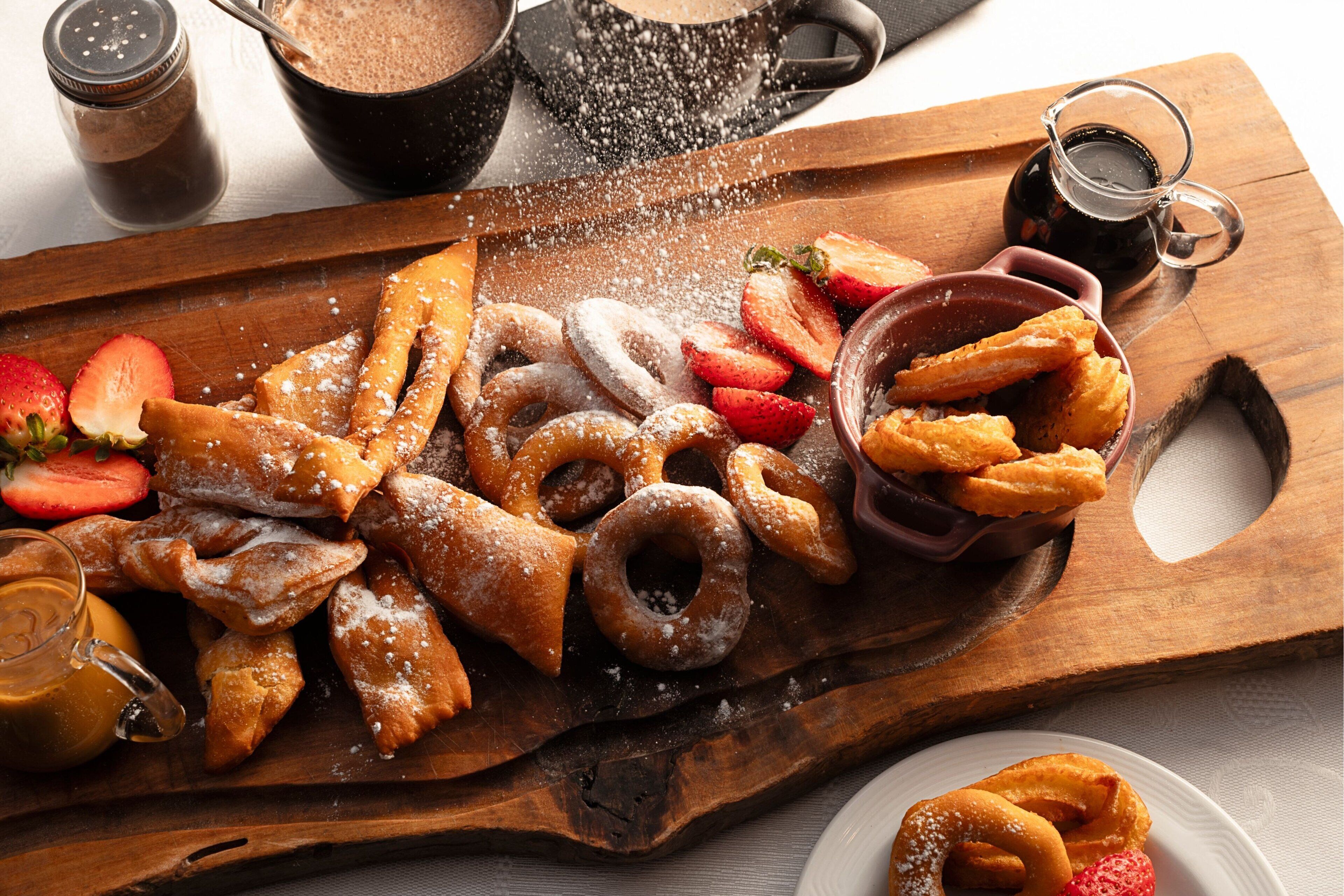 A wooden board with various pastries, including doughnuts and pretzels, dusted with powdered sugar, accompanied by strawberries and a glass of coffee.