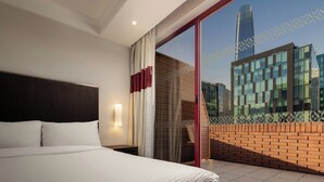 A hotel room with a large bed, a nightstand, a wall-mounted lamp, and a view of a cityscape through a glass door.