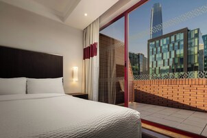 Habitación de hotel con una cama grande, una mesita de noche, una lámpara de pared y vista a un paisaje urbano a través de una puerta de cristal.