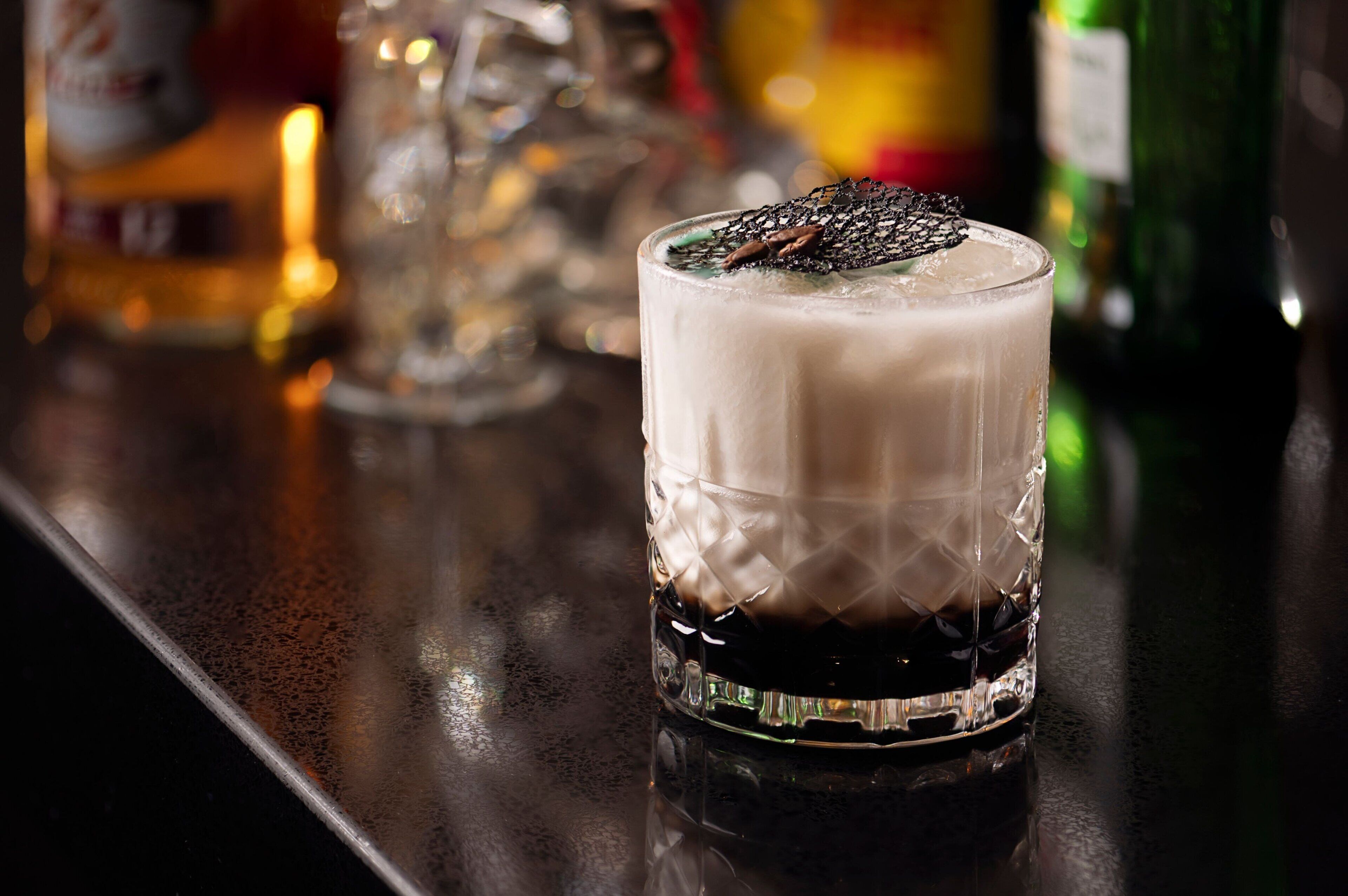 A glass of dark beverage with a frothy top, placed on a bar counter with various bottles and glasses in the background.