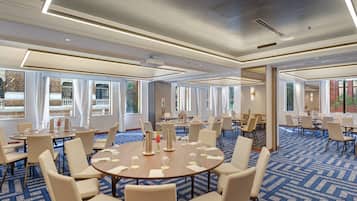 Un salón de conferencias de hotel con varias mesas y sillas, ventanas amplias y una alfombra estampada.
