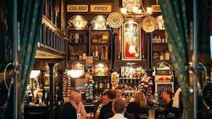 Un bar avec des étagères remplies de bouteilles, une horloge et des panneaux indiquant la présence de cigares et de pipes.