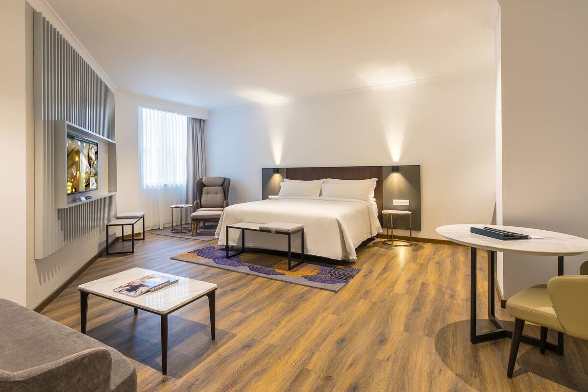 A hotel room with a bed, a desk, a chair, a small table, a bookshelf, and a window with curtains.