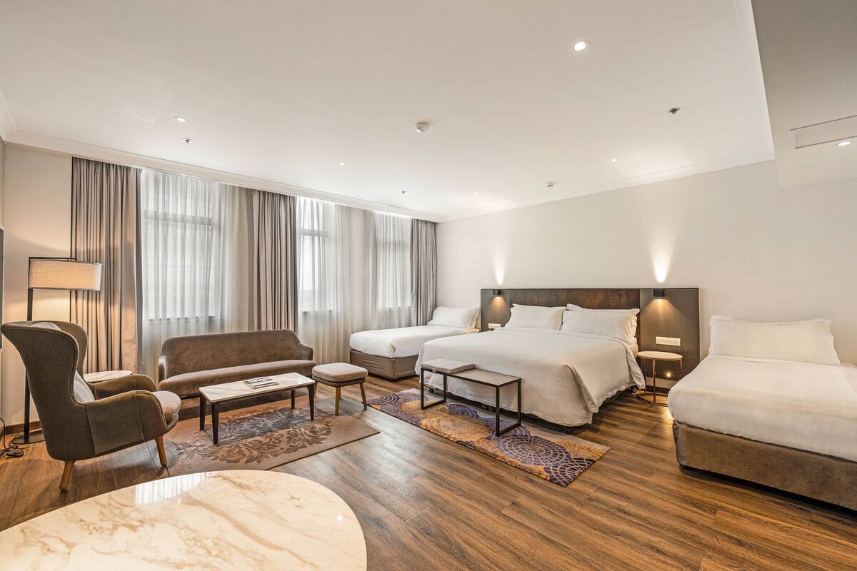 A modern hotel room with a large bed, two smaller beds, a sofa, a chair, a coffee table, and a lamp.