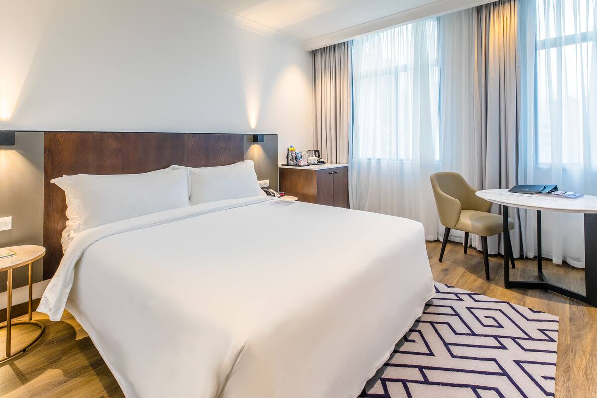 A hotel room with a large bed, a desk with a chair, a small table, and a nightstand.