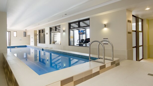 Une grande piscine intérieure avec une plage carrelée, un escalier en métal et une vue sur une salle de sport.