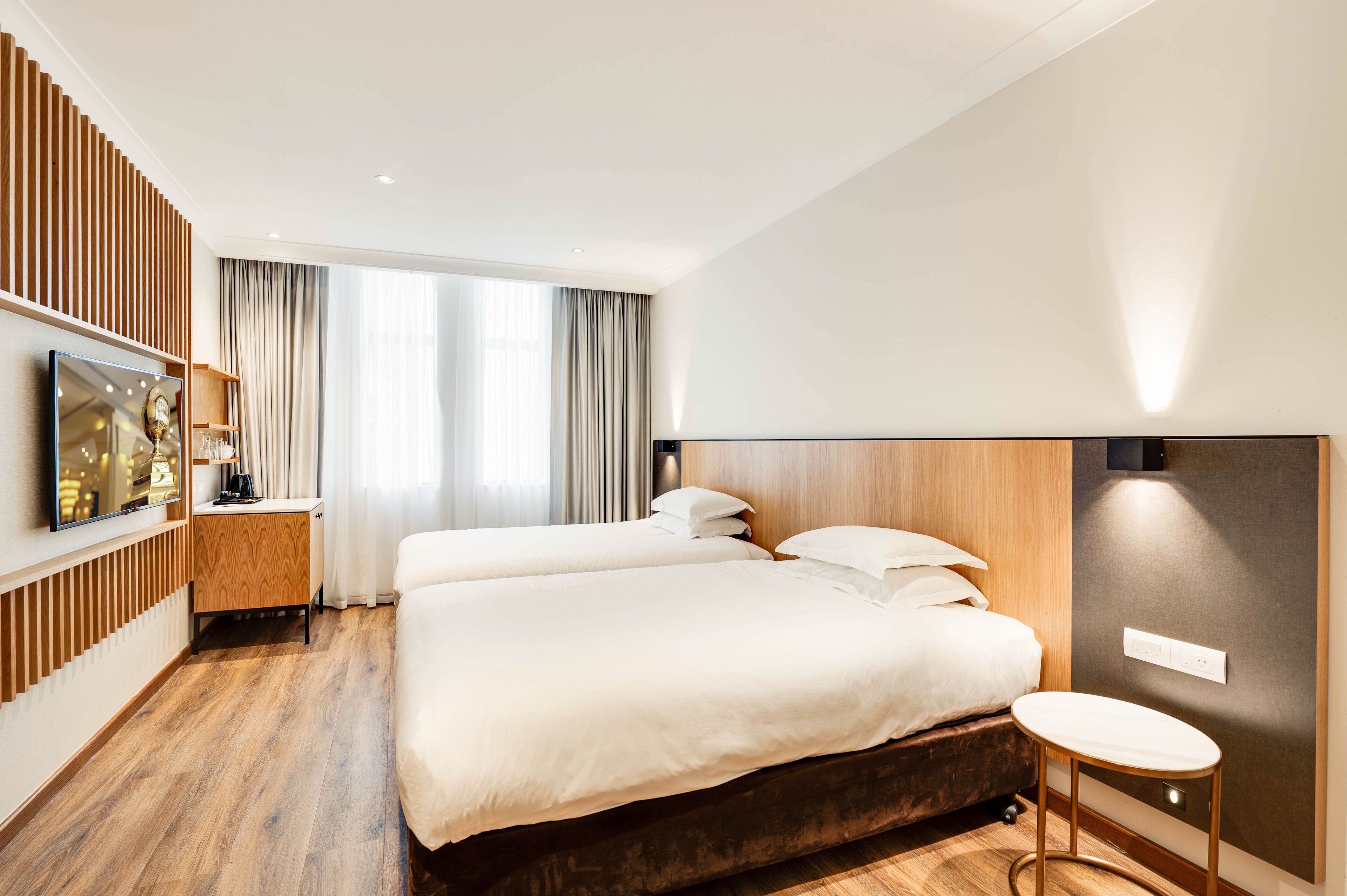 Una habitación de hotel moderna con dos camas, piso de madera, televisor sobre un soporte y un ventanal con cortinas.
