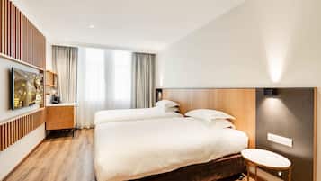 Una habitación de hotel moderna con dos camas, piso de madera, televisor sobre un soporte y un ventanal con cortinas.