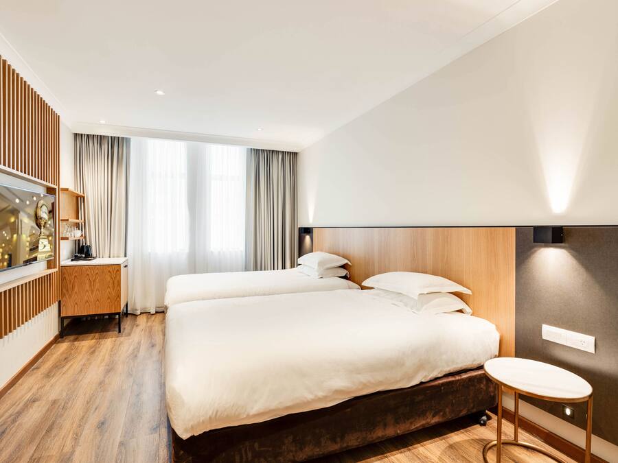 Una habitación de hotel moderna con dos camas, piso de madera, televisor sobre un soporte y un ventanal con cortinas.