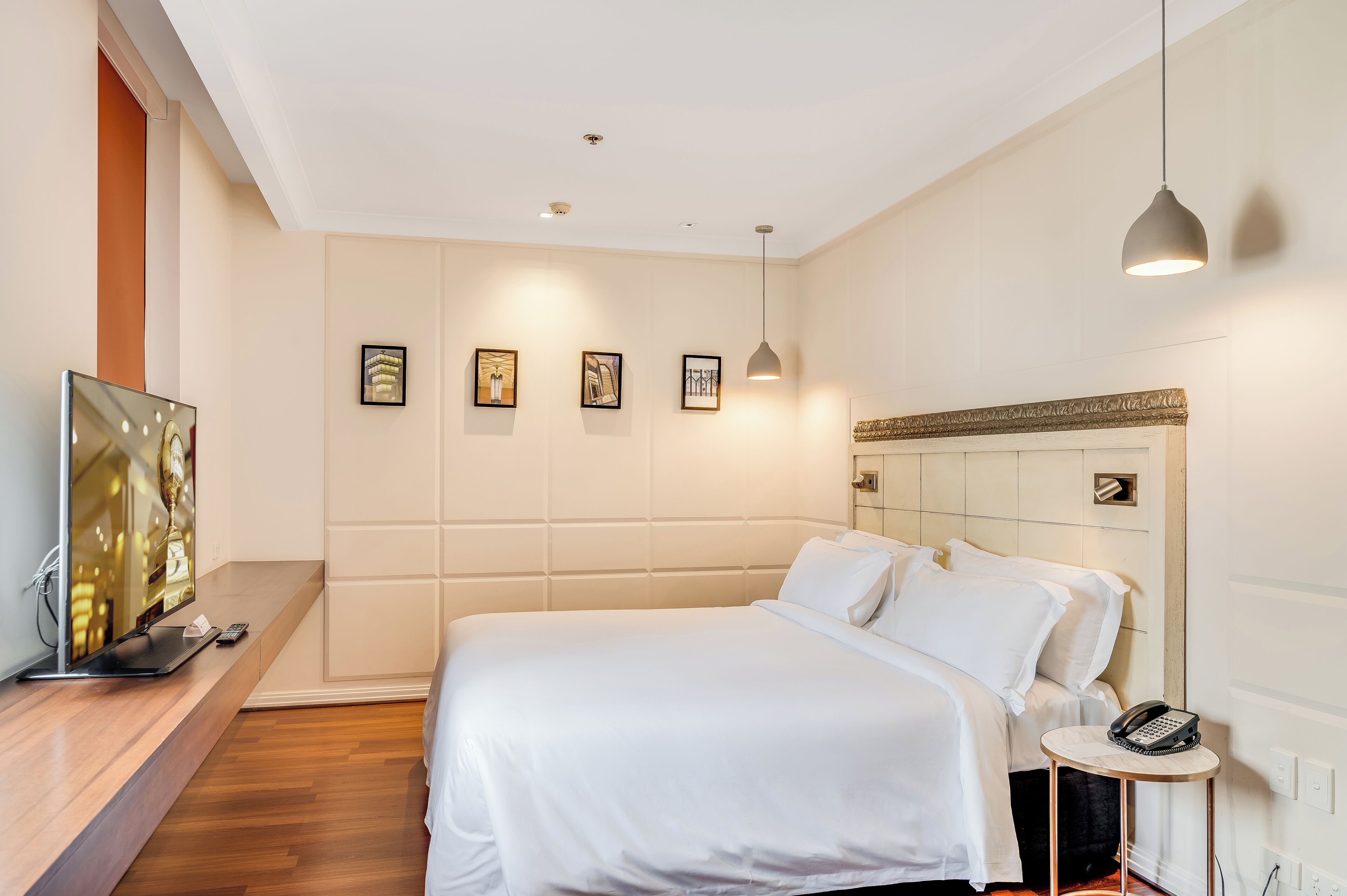 A modern hotel room with a large bed, a bedside table with a phone, a TV, and framed artwork on the wall.