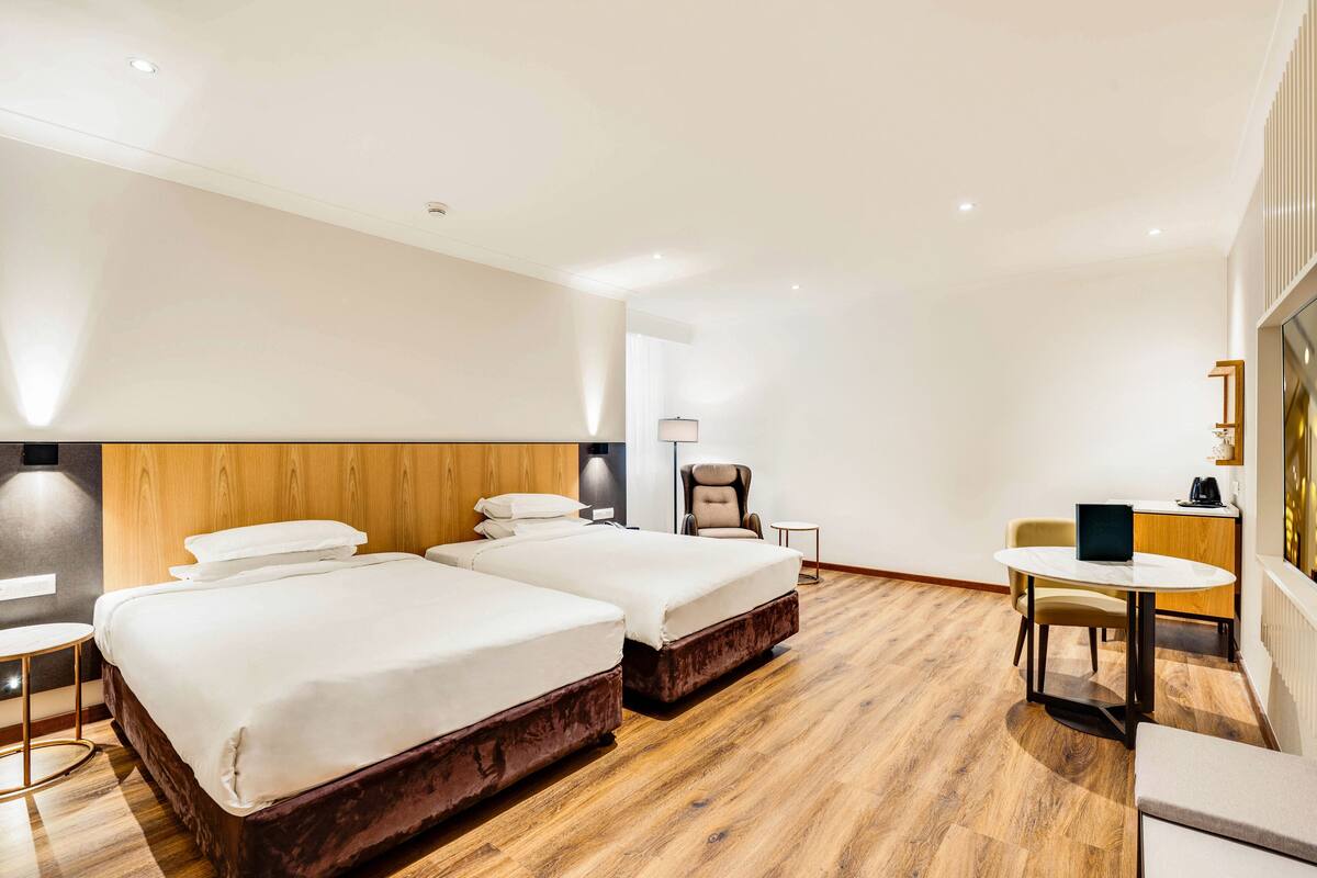 A hotel room with two beds, a desk, a chair, and a lamp.