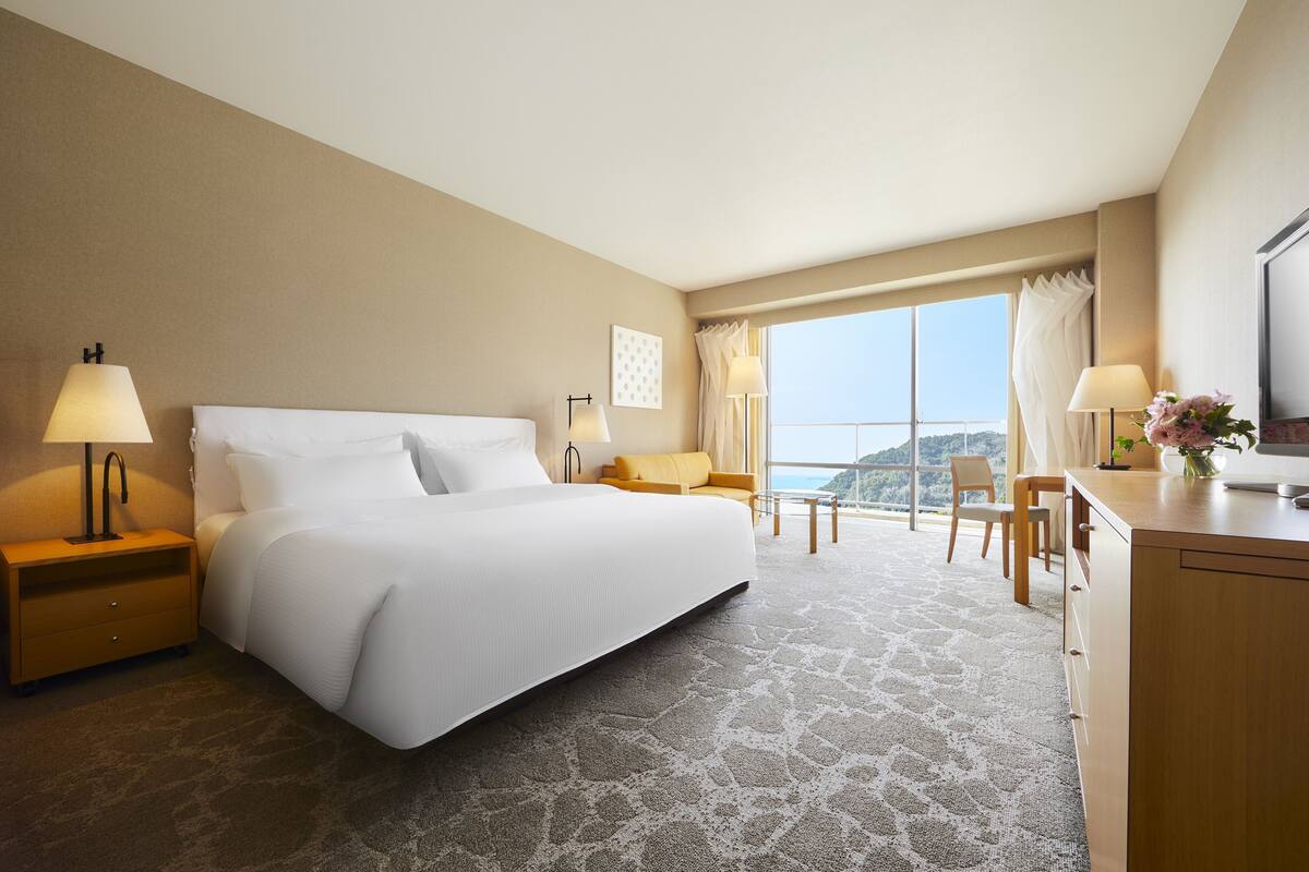 Habitación de hotel con una cama grande, dos mesitas de noche con lámparas, un escritorio de madera y vistas al exterior.