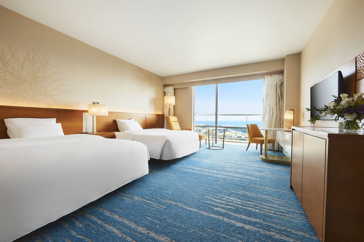 Habitación de hotel con dos camas, un escritorio, una silla y un ventanal con vistas.