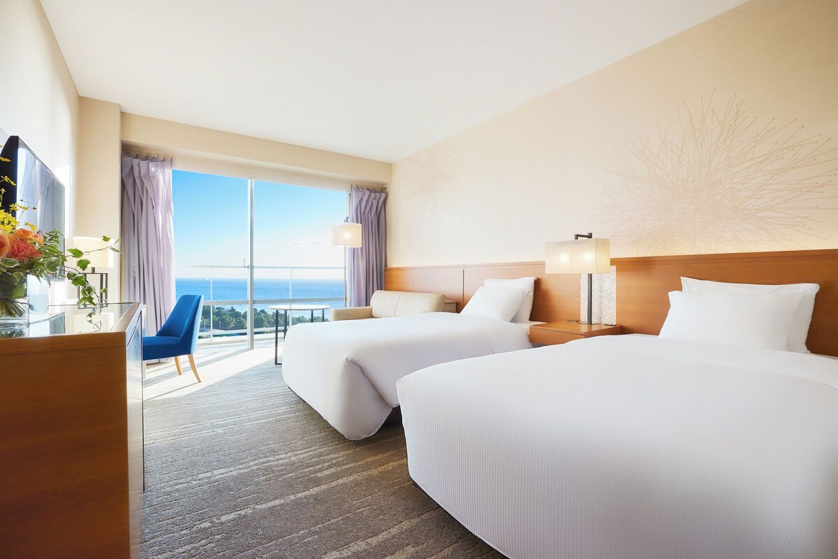 A hotel room with two beds, a desk with a lamp, a chair, and a view of the ocean.