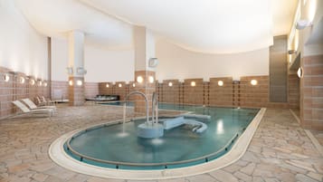 An indoor swimming pool with a stone floor, lounge chairs, and a changing room area.