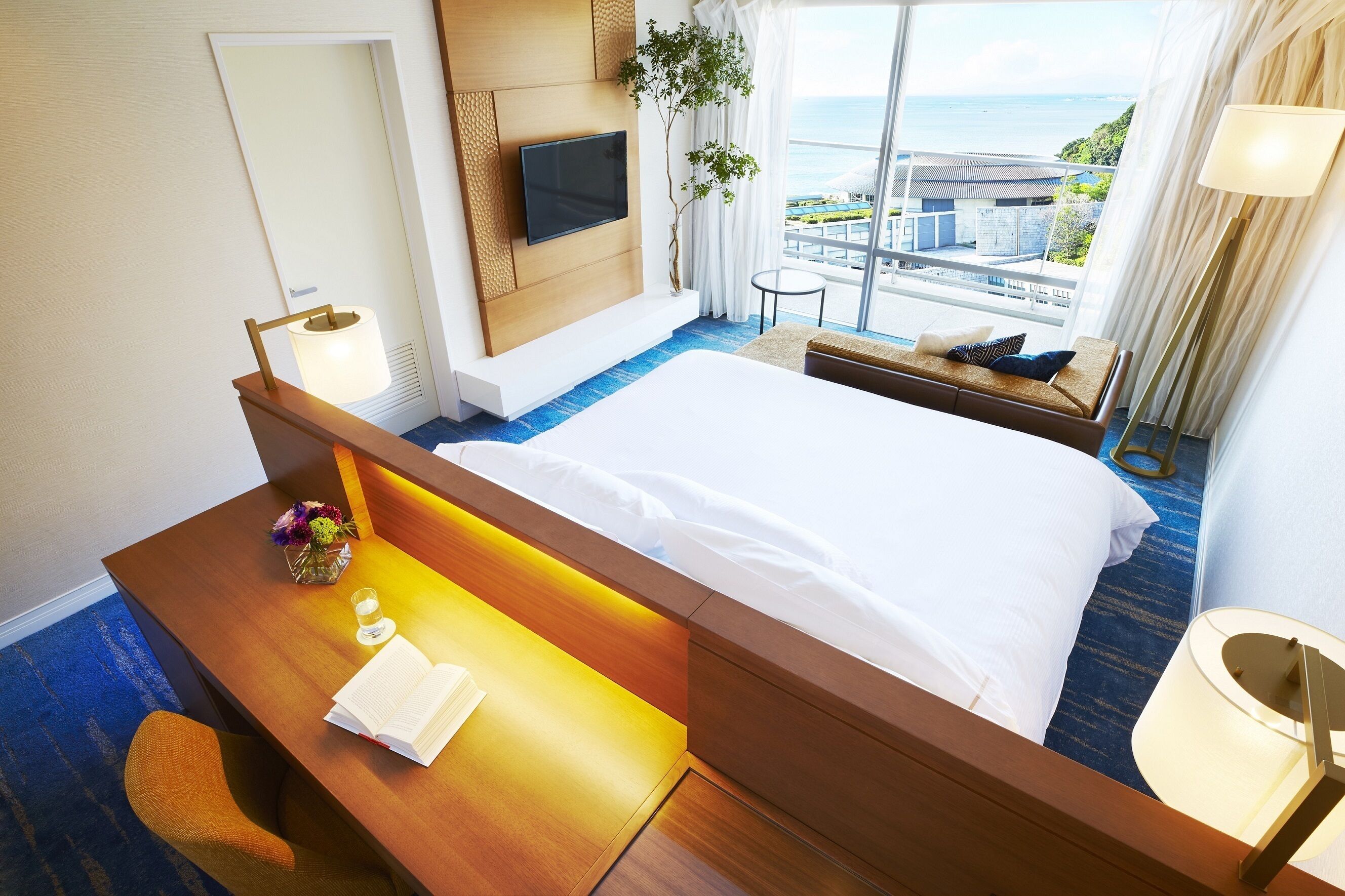 A hotel room with a large bed, a desk with a lamp, a TV, and a view of the ocean.