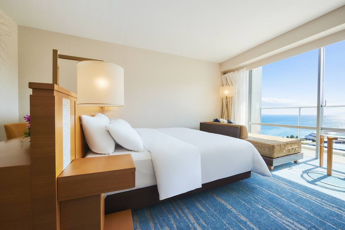 Habitación de hotel con una cama grande, una mesita de noche, una lámpara, una mesita auxiliar de madera y vistas al océano a través de un ventanal amplio.
