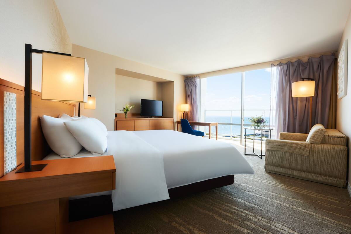 Habitación de hotel con una cama grande, un escritorio con televisor, una silla y vista al océano.