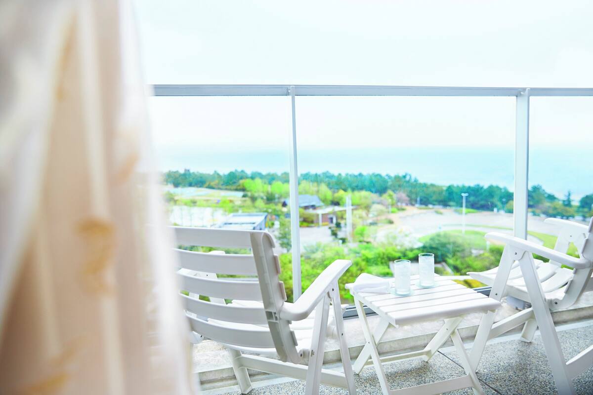 Un balcón con sillas y una mesa blancos, con vistas a un paisaje verde y una masa de agua.