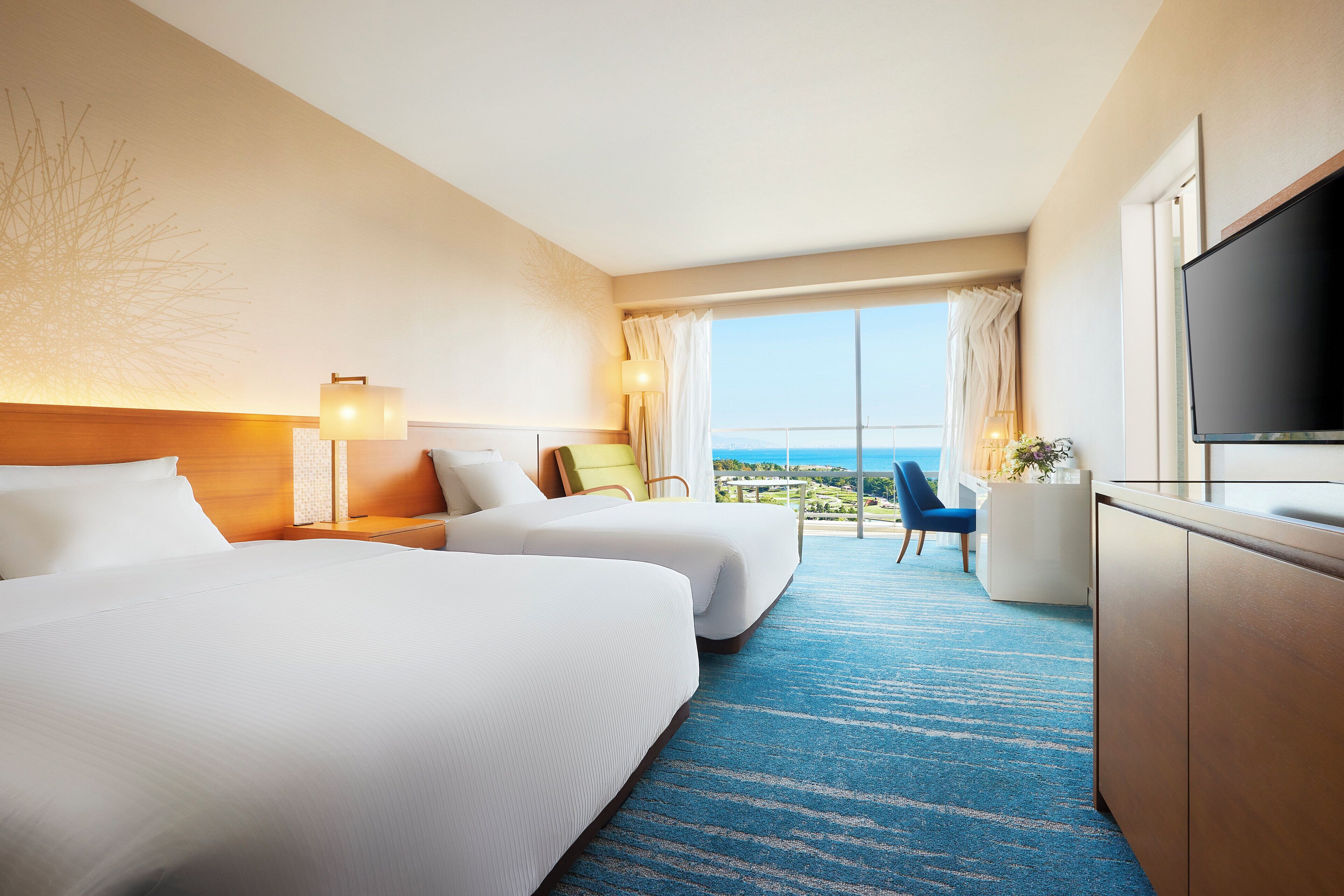 A hotel room with two beds, a TV, a desk, and a view of the ocean.