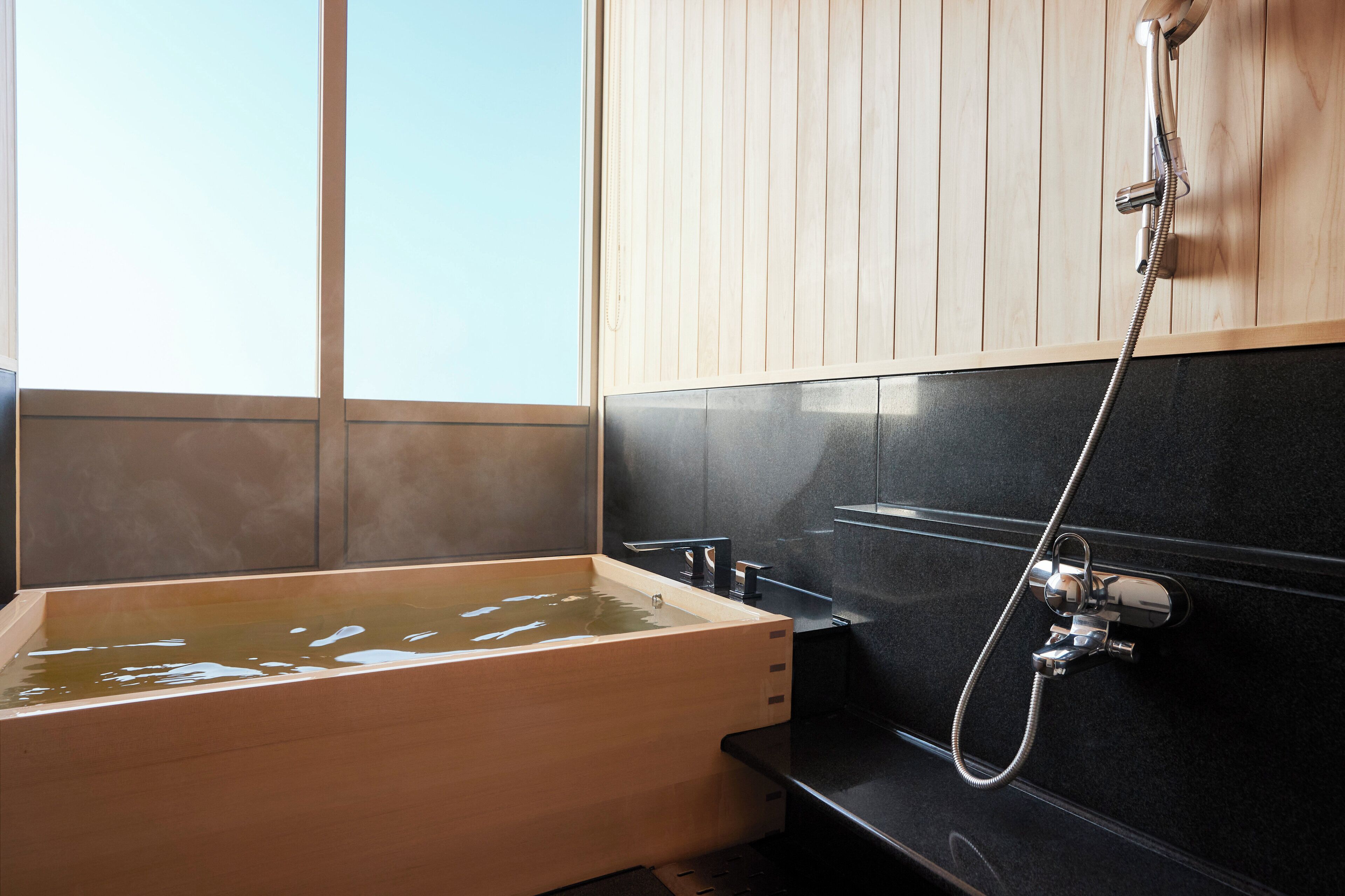 Une baignoire en bois avec une pomme de douche, un mur recouvert de carreaux noirs et un mur avec un panneau en bois.