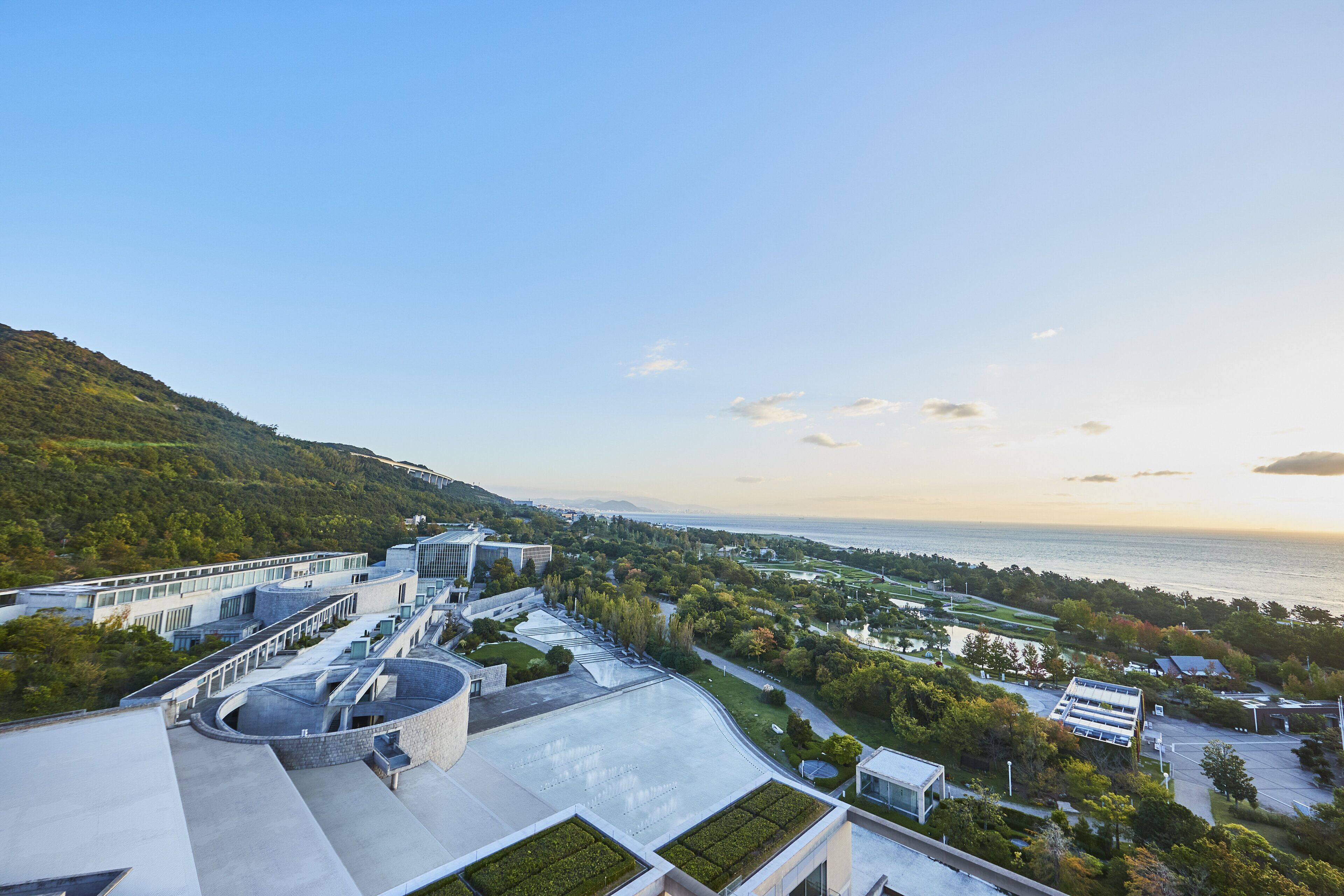 A modern building with a rooftop terrace, surrounded by greenery and a view of the sea.