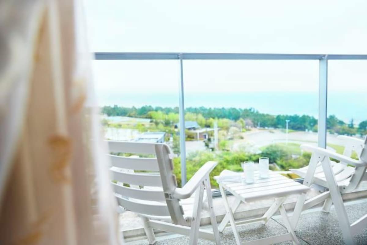 A balcony with white chairs and a table, overlooking a scenic landscape with greenery and a body of water.