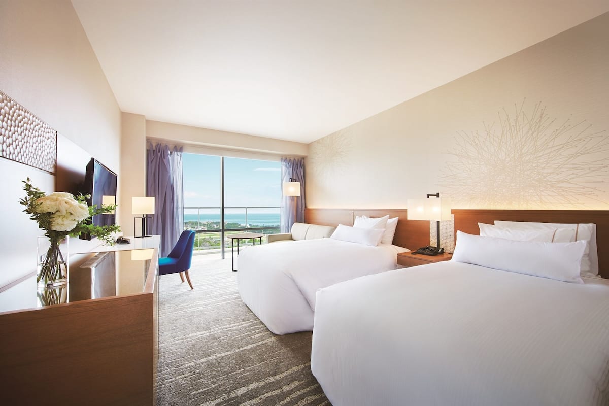 Habitación de hotel con dos camas, un escritorio con televisor, una mesita y vistas al exterior.