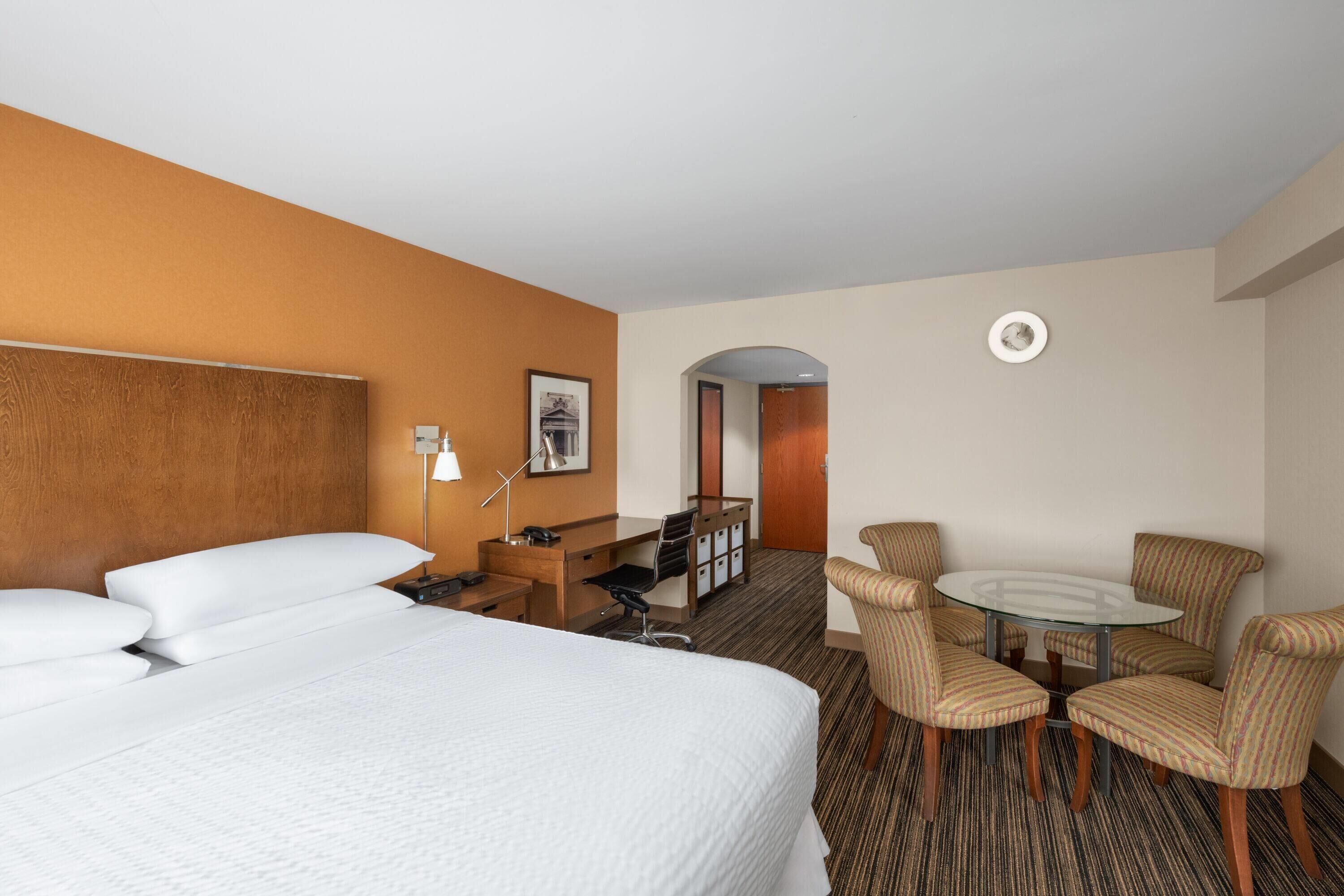 Une chambre d’hôtel avec un grand lit, un bureau avec une chaise, une petite table avec quatre chaises et une porte donnant sur une autre pièce.