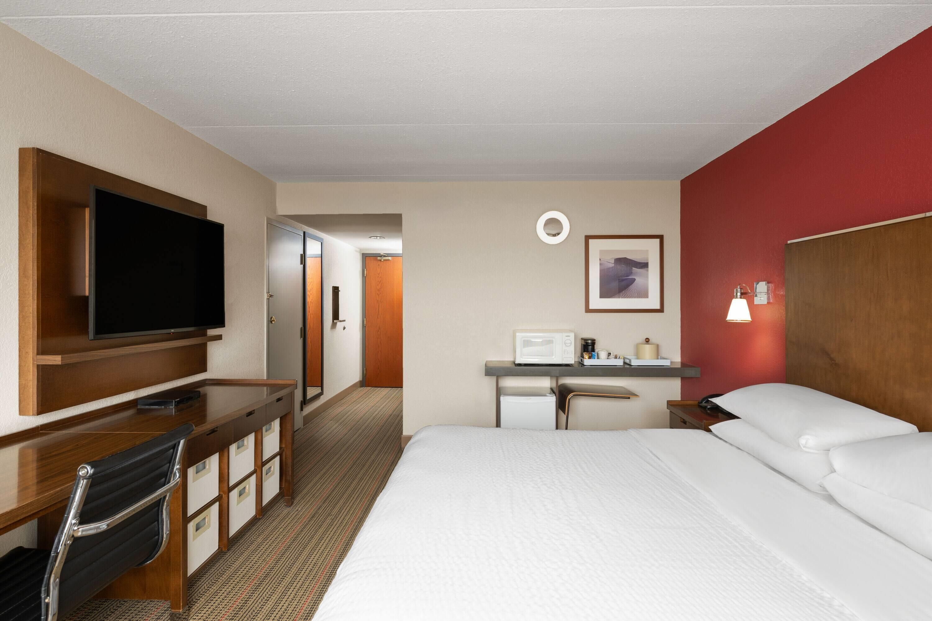 A hotel room with a bed, a desk with a television, a microwave, and a small table with a lamp.