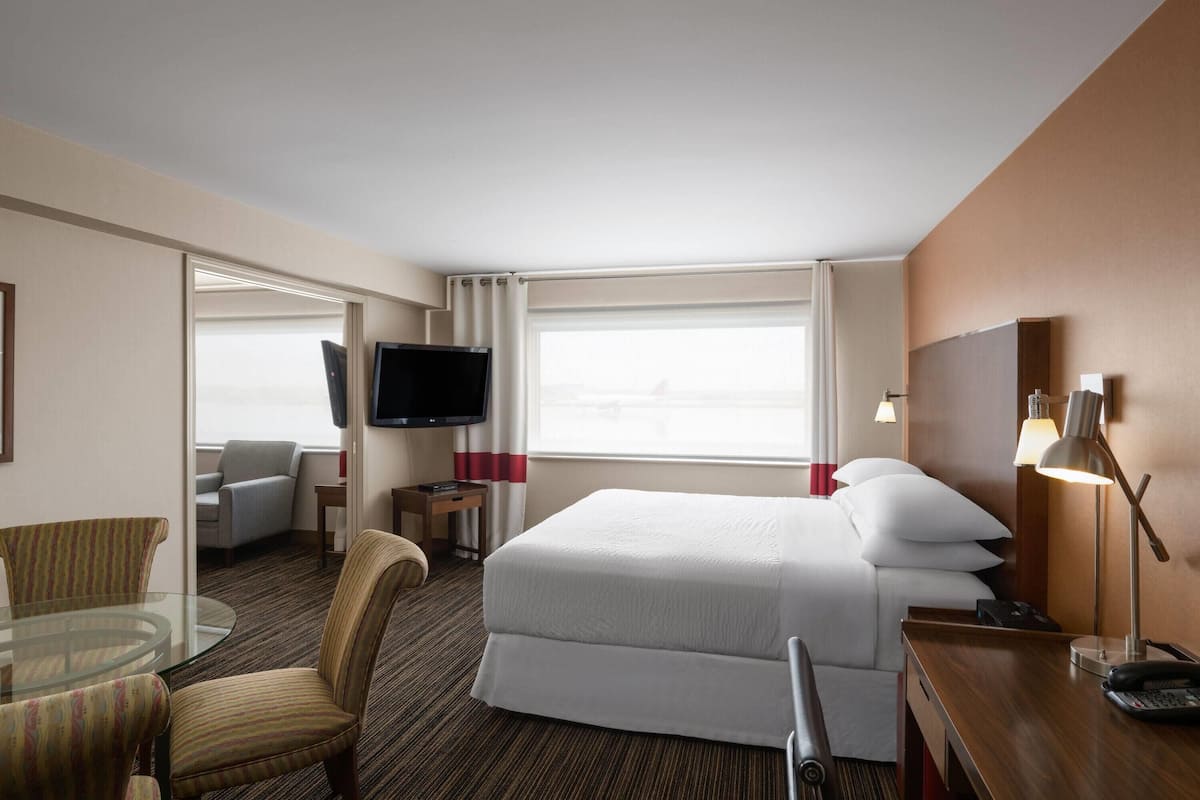 Habitación de hotel con una cama grande, un escritorio con lámpara, televisión, una mesa de vidrio con sillas y vistas al exterior a través de la ventana.