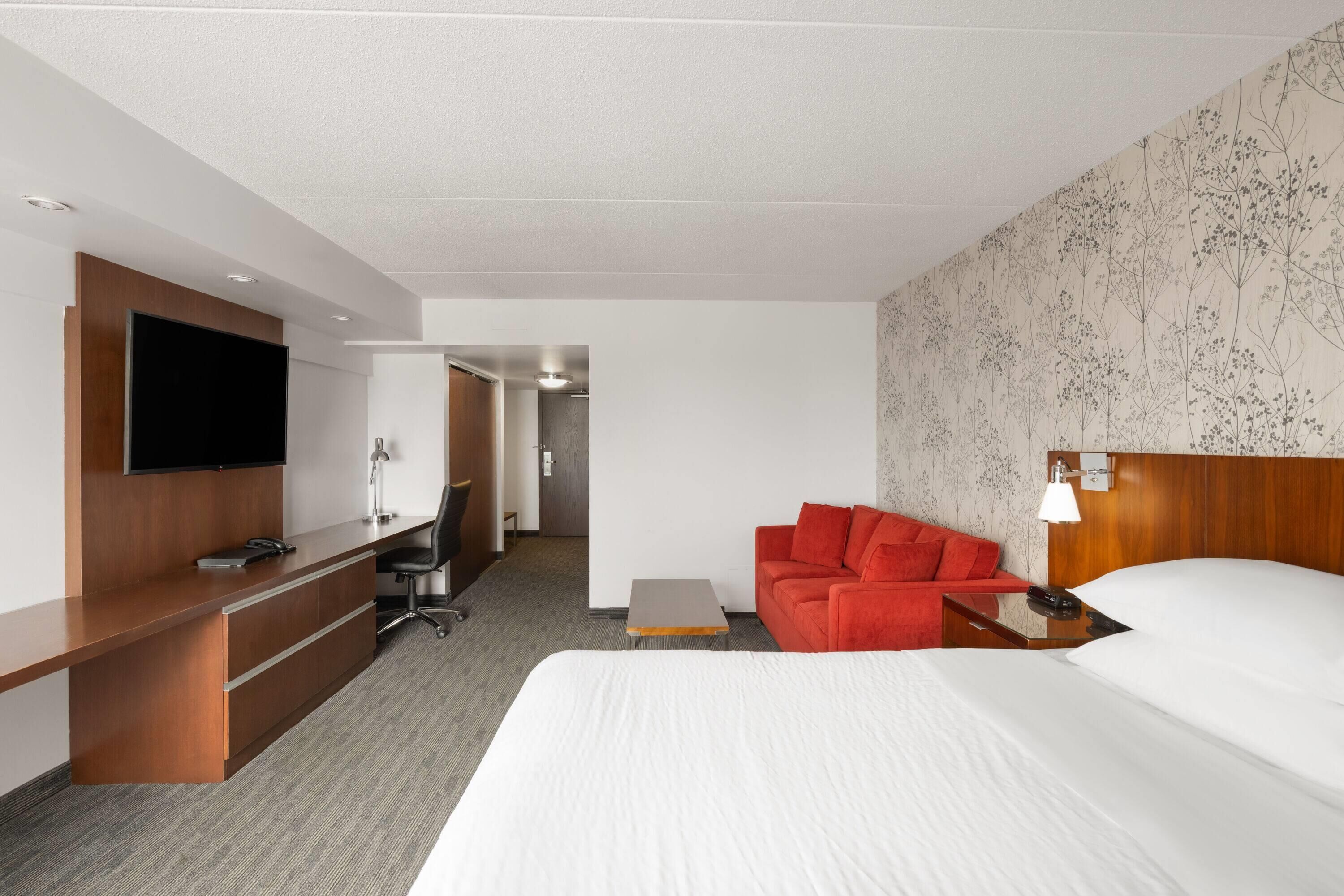 A modern hotel room with a large bed, a desk with a flat-screen TV, a red sofa, and a small table.