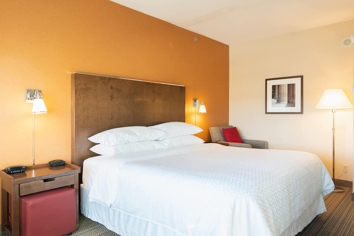 Una habitación de hotel con una cama grande, un cabecero de madera, una mesita de noche con lámpara, un banco rojo y un cuadro en la pared.