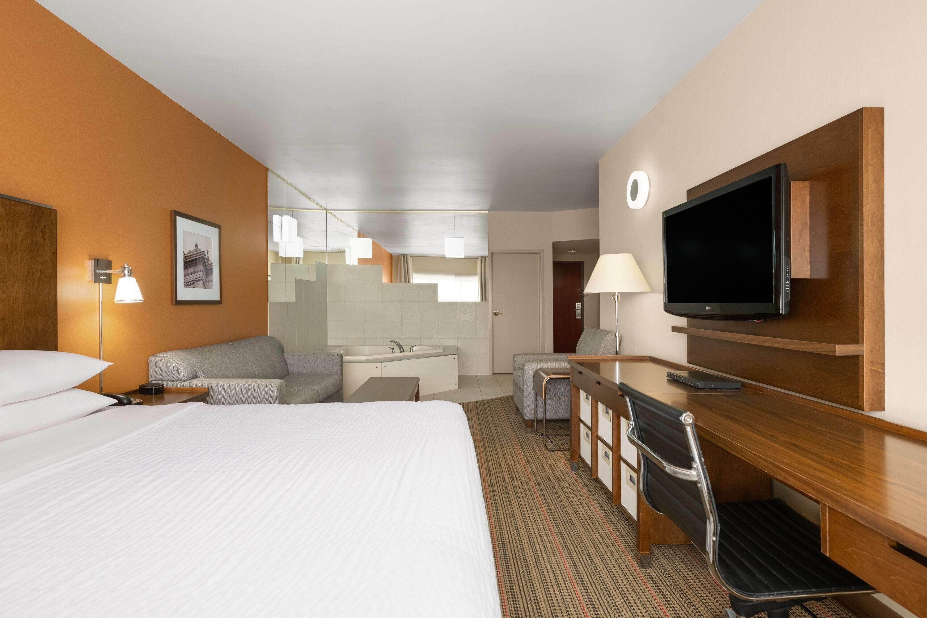 A hotel room with a bed, a desk with a chair, a television, a desk lamp, a sofa, and a bathroom with a sink and mirror.