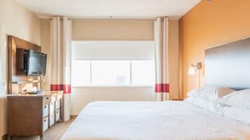 Quarto de hotel com uma cama grande, televisão na parede, escrivaninha com gavetas e janela com cortinas.