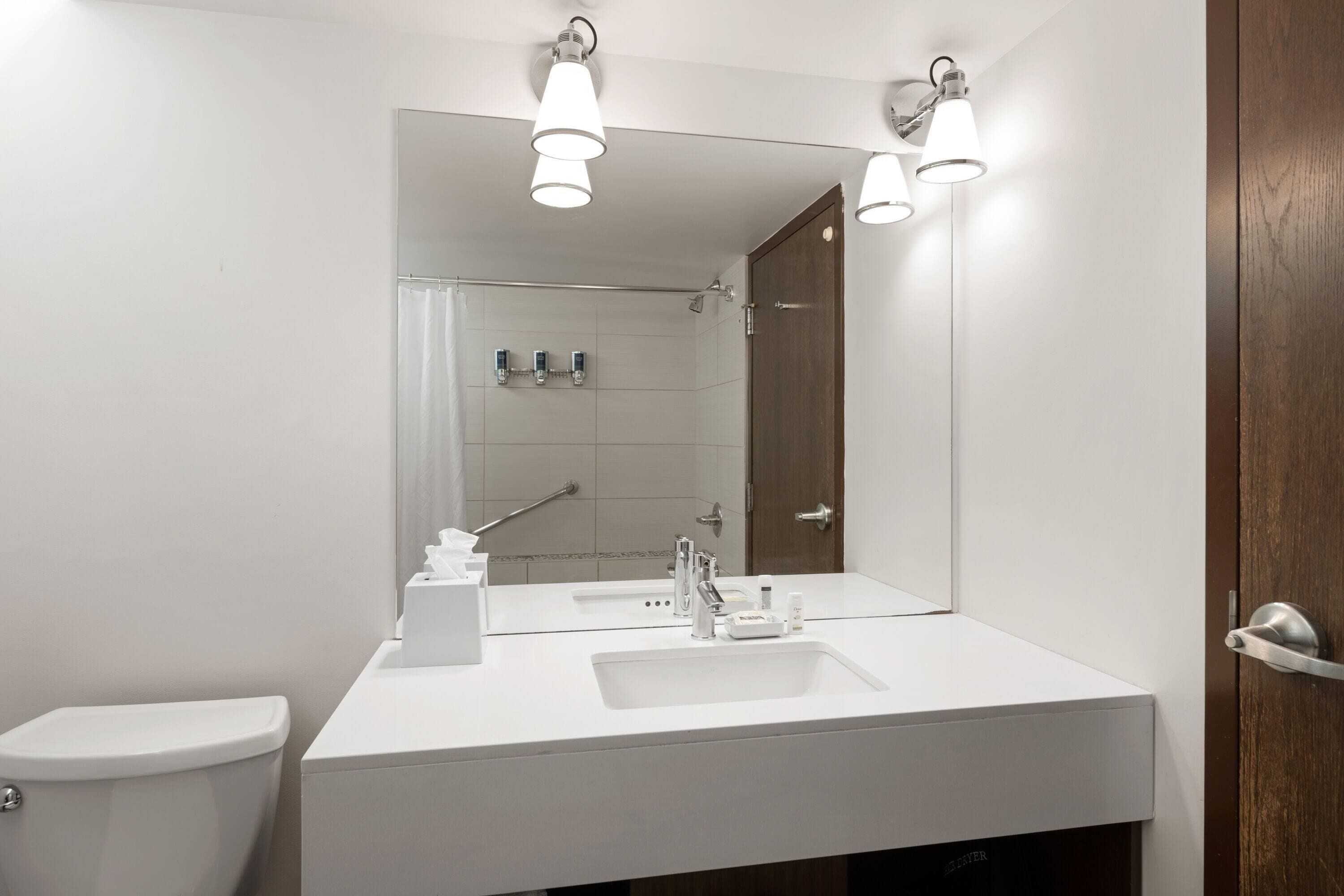 A modern bathroom with a white sink, mirror, and toilet. There are overhead lights and a shower visible in the background.
