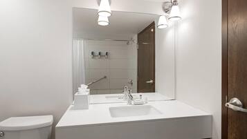 A modern bathroom with a white sink, mirror, and toilet. There are overhead lights and a shower visible in the background.