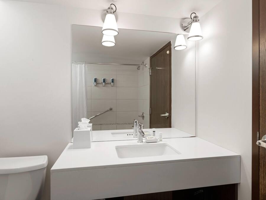 Un baño moderno con un lavamanos blanco, espejo y retrete. Se observan luces en el techo y una ducha al fondo.