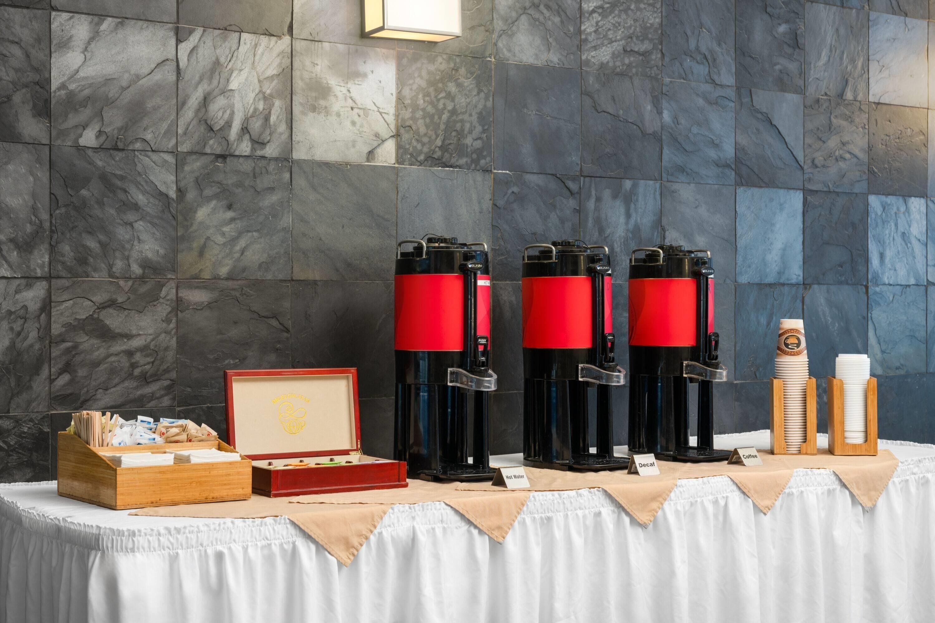 A coffee station with four red and black coffee machines, a wooden box with napkins, and a small display case with a nameplate.