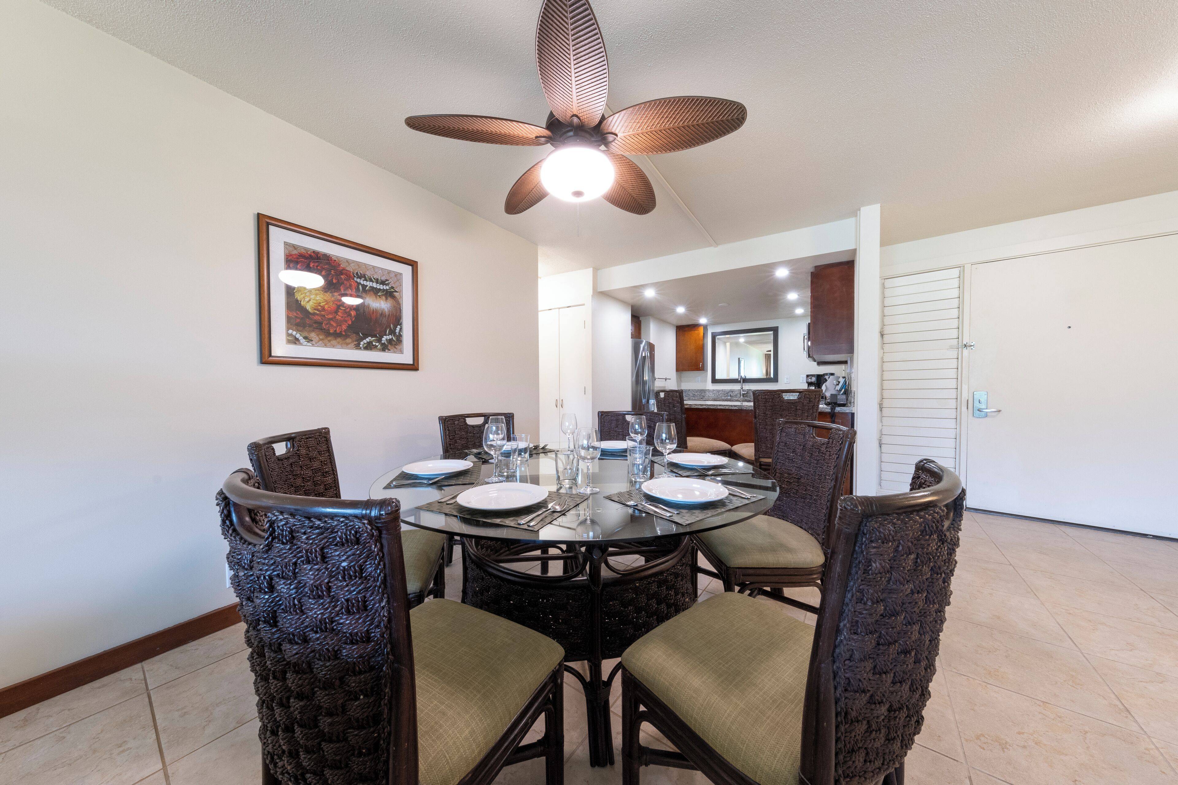 A dining area with a round table set for four, surrounded by wicker chairs with cushioned seats. A ceiling fan with a light fixture is above the table.
