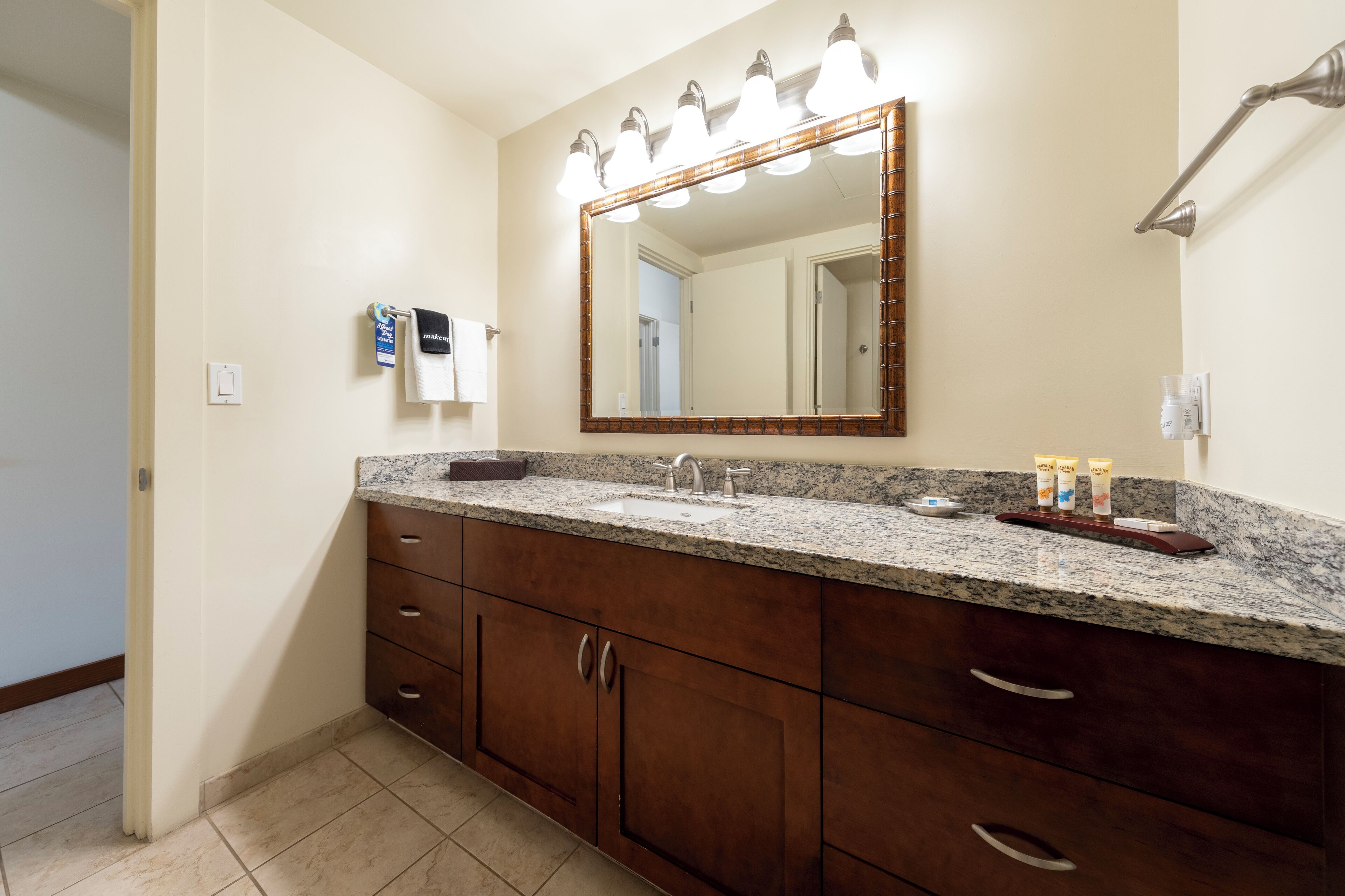 A bathroom with a granite countertop, a large mirror with lights, wooden cabinets, and toiletries.