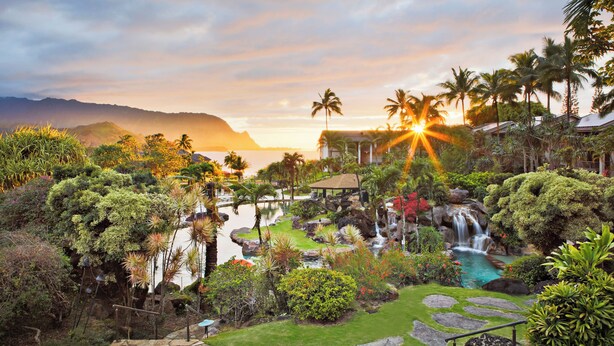 A resort with a waterfall, palm trees, and a view of the ocean at sunset. - Hanalei Bay Resort (Princeville)