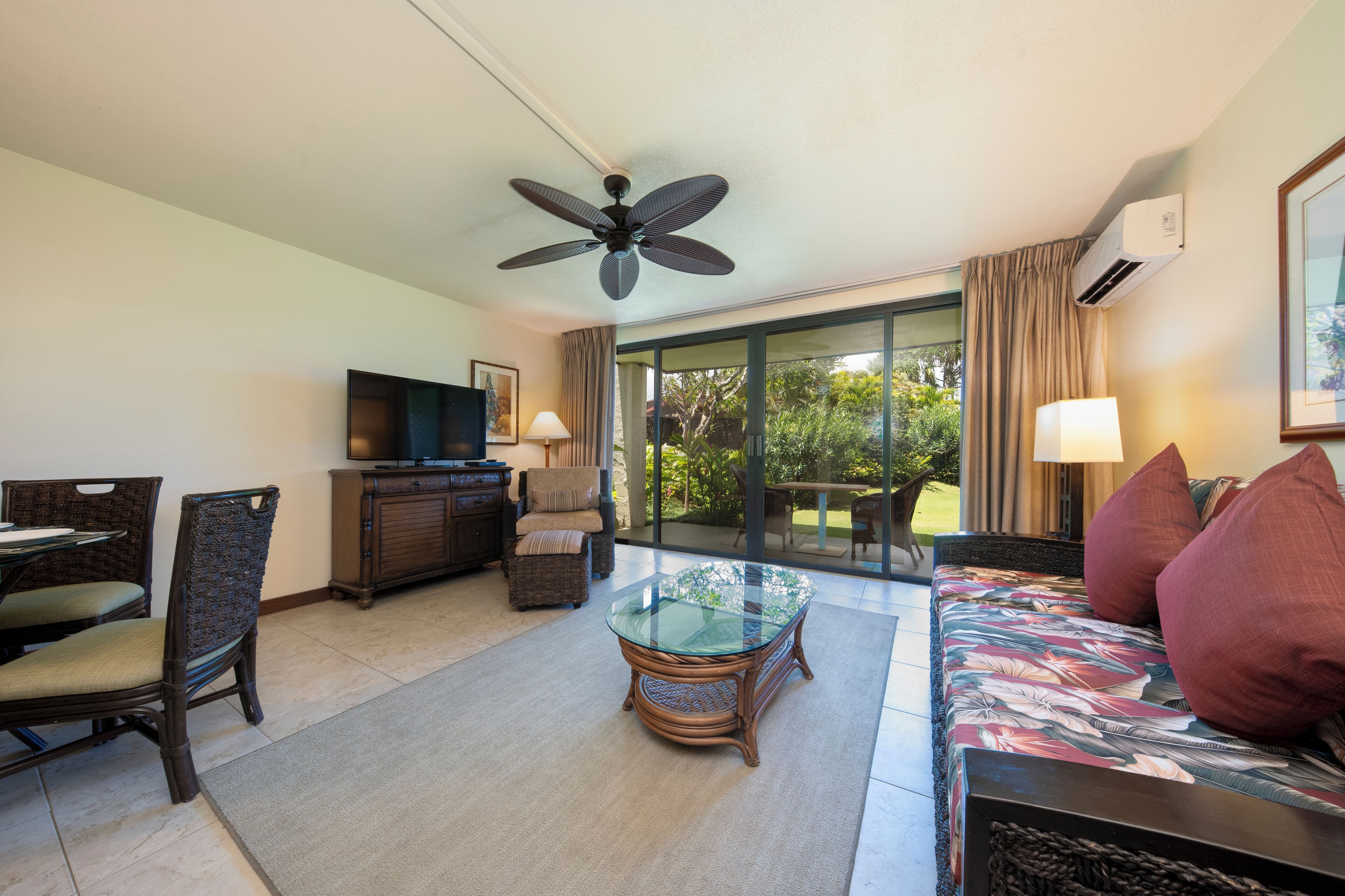 Habitación de hotel con comedor, sofá tapizado con motivos florales, mesa de centro de vidrio, sillas de mimbre, televisor, ventilador de techo y puerta corredera de vidrio que da a un jardín.