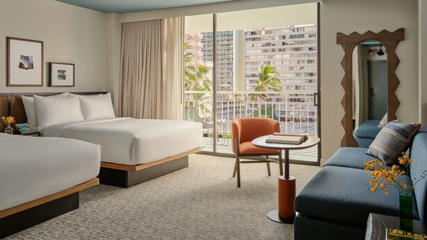 Habitación de hotel con dos camas, un sofá, una mesita y un balcón con vistas a edificios altos y palmeras.
