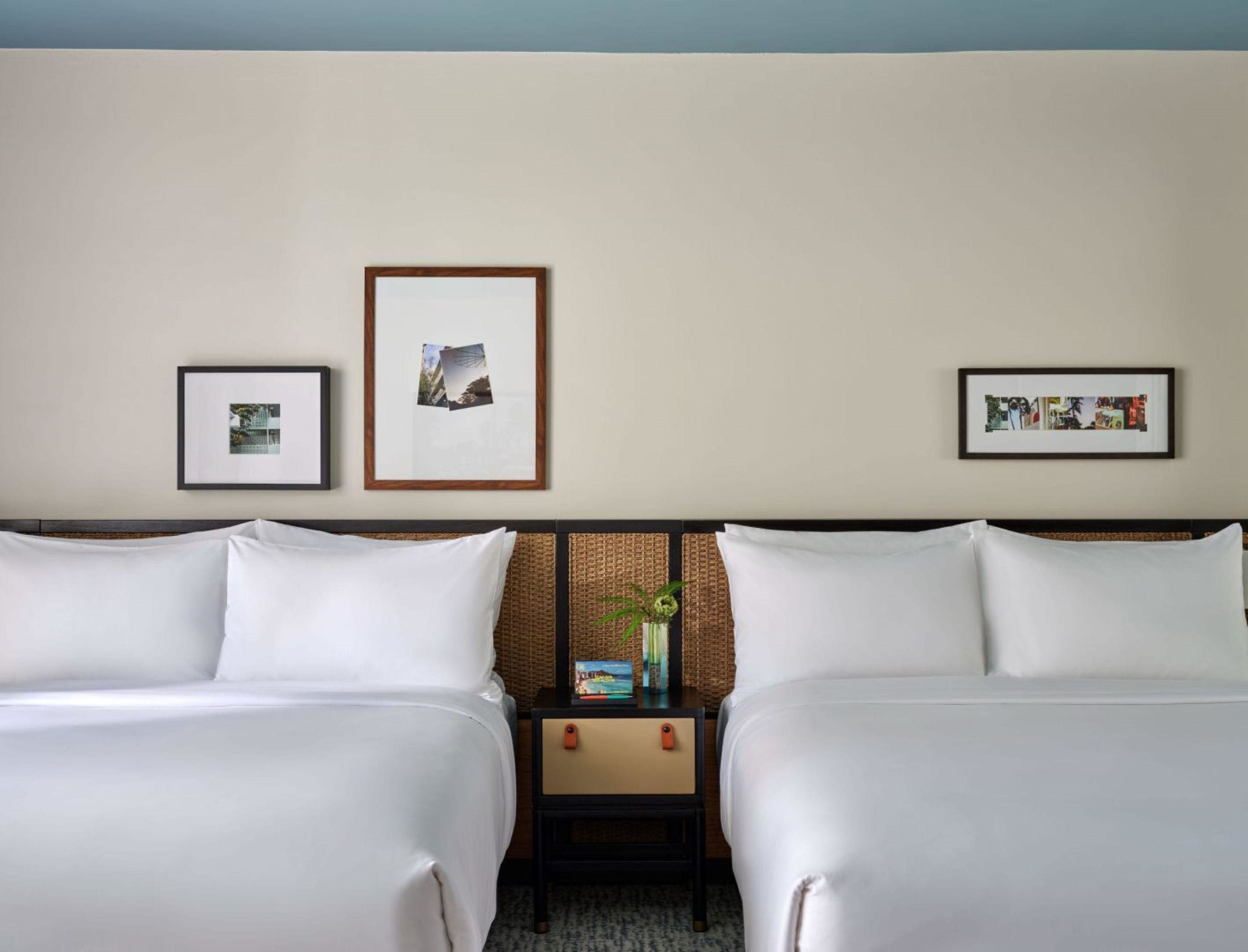 A hotel room with two beds, a nightstand with a vase, and three framed pictures on the wall.