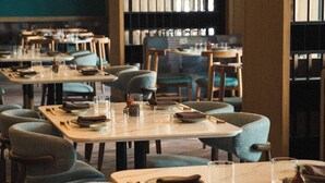 Un interior de restaurante con mesas dispuestas para comer, sillas, luces colgantes y estanterías al fondo.